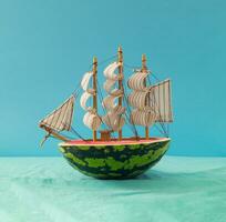 Watermelon sailboat traveling at the ocean. Sea ship made of watermelon against blue background. Minimal travel, vacation and summer journey concept. Creative food idea. Summer fruit aesthetic. photo