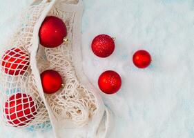 Creative composition made of red Christmas baubles coming out of mesh net bag on snowy background. Minimal Christmas or New Year concept. Trendy winter holidays idea. Flat lay, top of view. photo