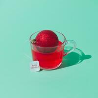 Creative Christmas composition of transparent glass cup of fruit tea with teabag made of red Christmas bauble on green background. Minimal Christmas or New Year concept. Trendy winter holidays idea. photo