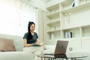 Asian woman sits on the sofa while taking notes from her computer screen while working or studying online at home. photo