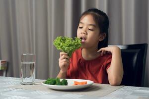 asiático pequeño niña comiendo sano vegetales con saborear. foto