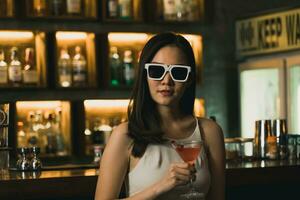 asiático mujer Bebiendo un cóctel a un bar mientras vistiendo Gafas de sol. foto