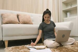 Asian woman sits on a rug in the living room and works on a computer while writing down notes in a notebook. photo