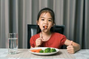 asiático pequeño niña comiendo sano vegetales con saborear. foto