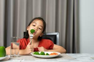 asiático pequeño niña comiendo sano vegetales con saborear. foto