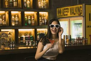 asiático mujer Bebiendo un cóctel a un bar mientras vistiendo Gafas de sol. foto