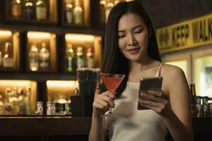 asiático mujer usado a charla con su amigos en Teléfono móvil mientras Bebiendo whisky a el bar. foto