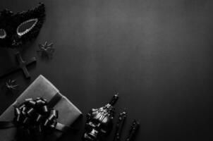 Víspera de Todos los Santos fiesta concepto con negro accesorios, regalo caja, lujoso mascarilla, el cruzar y cráneo vela en negro antecedentes. foto