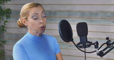 A girl in a blue turtleneck, blonde, hosts a podcast. Large microf on a stand against the background of a ficus video