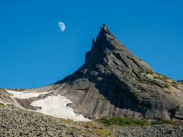 Pointed cliff with a small snow glacier, moon in the blue evening sky. Awesome scenic mountain landscape with big pointed stones in evening. Sharp rocks background. Western Sayans. photo