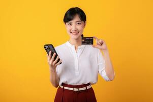 Portrait beautiful young asian woman enterpriser happy smile wearing white shirt and red plants holding credit card and smartphone isolation on yellow background. Online shopping concept. photo