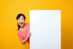 young asian sports fitness woman happy smile wearing pink sportswear standing behind the white blank banner or empty space advertisement board against yellow background. wellbeing lifestyle concept. photo