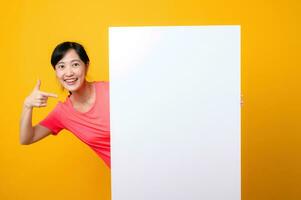 young asian sports fitness woman happy smile wearing pink sportswear standing behind the white blank banner or empty space advertisement board against yellow background. wellbeing lifestyle concept. photo