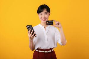 Portrait beautiful young asian woman enterpriser happy smile wearing white shirt and red plants holding credit card and smartphone isolation on yellow background. Online shopping concept. photo