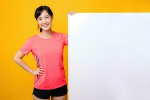 young asian sports fitness woman happy smile wearing pink sportswear standing behind the white blank banner or empty space advertisement board against yellow background. wellbeing lifestyle concept. photo