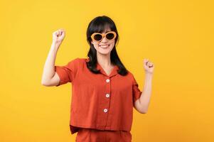 Portrait young beautiful asian woman happy smile dressed in orange clothes showing fist up hand gesture isolated on yellow studio background. Successful hooray celebrate young person concept. photo