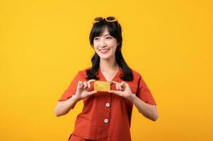 Portrait young beautiful asian woman happy smile dressed in orange clothes showing credit card isolated on yellow background. Pay and purchase shopping payment concept. photo