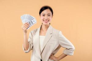 portrait of happy successful confident young asian business woman wearing white jacket holding cash money dollars standing over beige background. millionaire business, shopping concept. photo