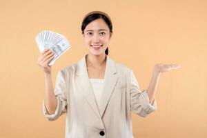 portrait of happy successful confident young asian business woman wearing white jacket holding cash money dollars standing over beige background. millionaire business, shopping concept. photo
