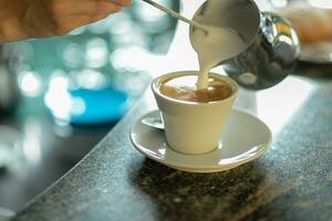 manos de joven mujer barista torrencial al vapor cremoso Leche en capuchino taza a el bar encimera. foto