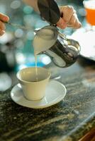 manos de joven mujer barista torrencial al vapor cremoso Leche en capuchino taza a el bar encimera. foto