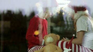 two kids in pajamas sitting on a wooden floor near the window with christmas light video