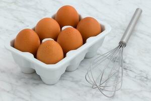 Eggs in a bowl and whisk on a white marble background photo