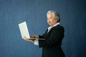 Portrait of intelligent senior businesswoman CEO manager holding in hands laptop creating a presentation on isolated blue background. Old Woman thinking after getting financial statements reports. photo