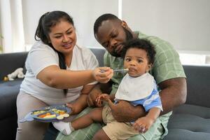 Asian mother feeding her 9 months old her cute little baby and African American helping for holding food plate At Home. Photo series of family, kids and happy people concept. Parents feed kids.