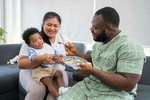 asiático madre alimentación su 9 9 meses antiguo su linda pequeño bebé y africano americano Ayudar para participación comida plato a hogar. foto serie de familia, niños y contento personas concepto. padres alimentar niños.