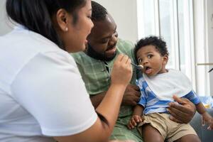 Asian mother feeding her 9 months old her cute little baby and African American helping for holding food plate At Home. Photo series of family, kids and happy people concept. Parents feed kids.