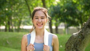 retrato joven asiático mujer atractivo sonriente y utilizar blanco toalla descansando después ejercicio. sonriente deportivo joven mujer trabajando fuera al aire libre y mirando a cámara. sano estilo de vida bien siendo bienestar foto