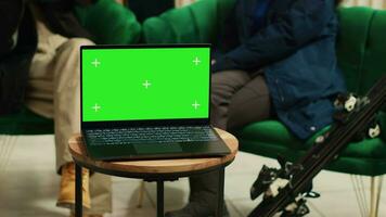 Digital device with greenscreen template placed on coffee table in lounge area at luxury alpine ski resort. People sitting on couch chatting, laptop running isolated copyspace display at hotel. video