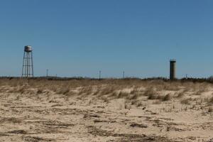 Sunset beach in Cape May New Jersey. The sand is a pretty brown. The dunes showing green tall grass to help prevent erosion. There is a lookout tower in the distance as well as a water tower. photo