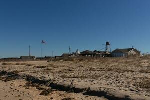 This is an image of the shops at Sunset beach in Cape May New Jersey. The beautiful sand leading up to it with green dunes gives color to this. The American flag is flying proudly by the buildings. photo