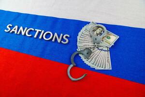 the word sanctions laid with silver metal letters on russian tricolor flag near dollar banknotes and handcuffs in diagonal perspective photo