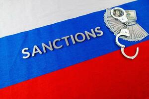 the word sanctions laid with silver metal letters on russian tricolor flag near dollar banknotes and handcuffs in diagonal perspective photo