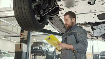 Low angle shot of a bearded car mechanic taking notes, examining lifted auto video