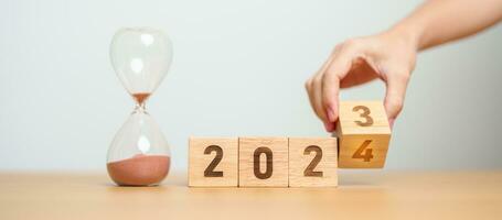 hand flipping block 2023 to 2024 text with hourglass on table. Resolution, time, plan, goal, motivation, reboot, countdown  and New Year holiday concepts photo