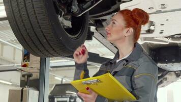 Female car mechanic examining wheels of a car on a lift, taking notes on clipboard video