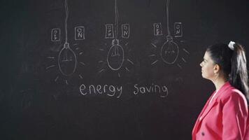 Woman writing energy saving on blackboard smiling looking at camera. video