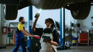 gekwalificeerd technicus staand onder geschorst auto in garage werkruimte, gebruik makend van werk licht naar controleren voor schade en tablet naar bestellen nieuw vervangen onderdelen na vinden problemen gedurende routine- controle video