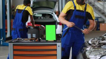 Panning shot of chroma key tablet in garage workspace sitting on work bench. Mockup device in car service next to professional tools with repairmen in background refurbishing damaged vehicles video