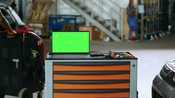 Grün Bildschirm Laptop platziert auf Arbeiten Bank im beschäftigt Garage Nächster zu Fachmann Werkzeuge während Angestellte durchstreifen um im verschwommen Hintergrund. Chroma Schlüssel Gerät im Auto Reparatur Geschäft video