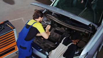 Repair shop employees using torque wrench and pliers to fix car in need of repairments. Coworkers refurbishing busted vehicle, checking for oil leakage using professional tools video