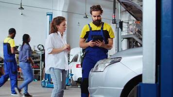 monteur luisteren naar klant in auto reparatie winkel, gebruik makend van tablet naar schrijven naar beneden auto wijzigingen ze wil naar maken. deskundige in garage met cliënt, luisteren naar haar verzoeken voor voertuig afstemmen video