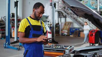 retrato de sonriente bipoc mecánico en garaje utilizando tableta a seguir Lista de Verificación mientras haciendo mantenimiento en cliente coche. alegre especialista en auto reparar tienda lo hace chequeo en vehículo video