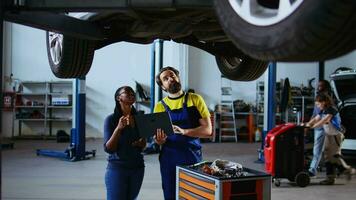 tecnico addetto alla manutenzione e cliente in piedi sotto auto nel box auto utilizzando il computer portatile per ordine nuovo parti dopo scoperta danni. dipendente assistere cliente di guardare in linea per sostituzione automobile componenti video