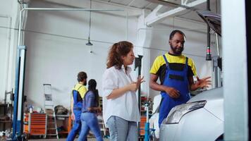 Experienced serviceman in car service showing customer what needs to be changed on her automobile for it to properly work. Garage professional inspecting vehicle placed on lift, chatting with client video