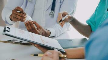 Multiracial medical team having a meeting with doctors in white lab coats and surgical scrubs seated at a table discussing a patients records video
